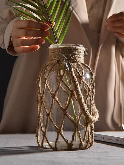 A hand‑woven hemp rope glass vase — a clear glass vessel wrapped in natural hemp fiber. Perfect for dried flowers, it adds texture and warm, rustic charm. Ideal for boho, Nordic, or minimalist interiors.